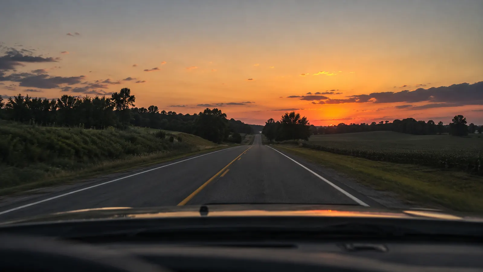 A quiet two-lane highway ahead of an older SUV at dusk, open road leading toward a hazy horizon.